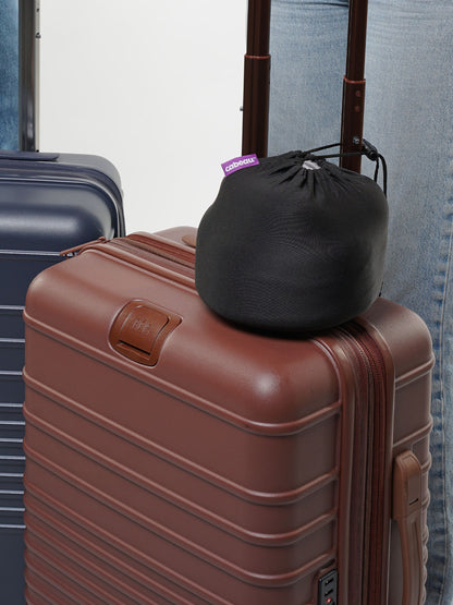 Brown suitcase with a black drawstring  travel bag with a neck pillow inside attached to the luggage, against a neutral background.