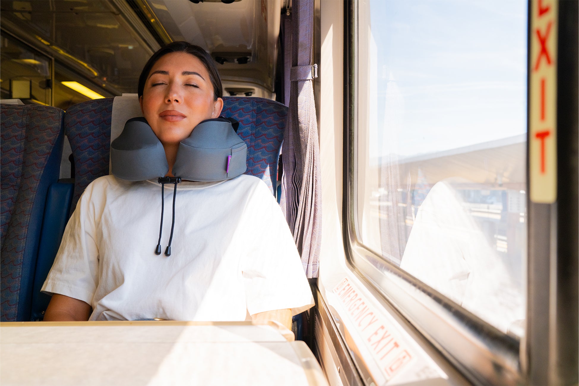 A woman rests her head on a gray neck pillow while seated in a train carriage next to a window, wearing a white t-shirt.